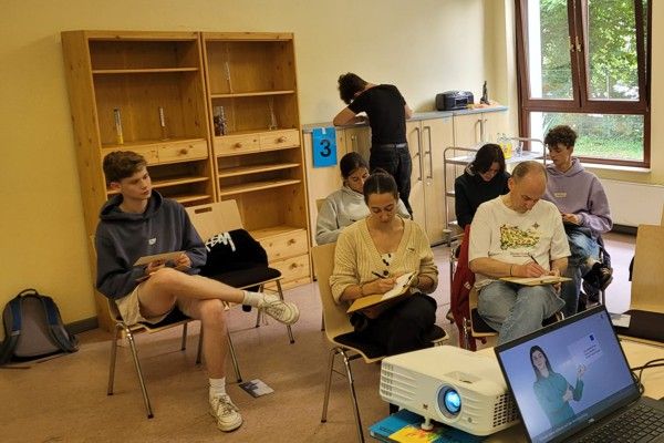 Junge Erwachsene sitzen in einem Seminarraum, arbeiten konzentriert an einer Aufgabe, während im Vordergrund ein Beamer läuft.