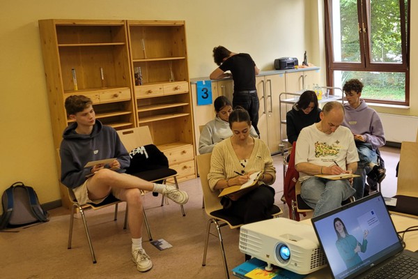 Junge Erwachsene sitzen in einem Seminarraum, arbeiten konzentriert an einer Aufgabe, während im Vordergrund ein Beamer läuft.